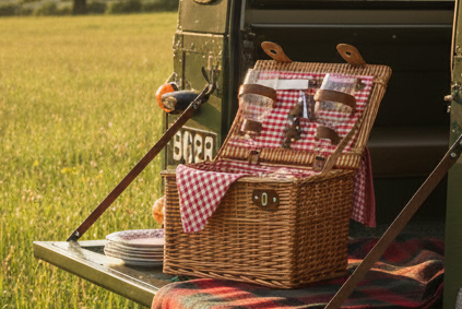 Wicker picnic basket with checkered cloth on the back of a green Land Rover in a field.